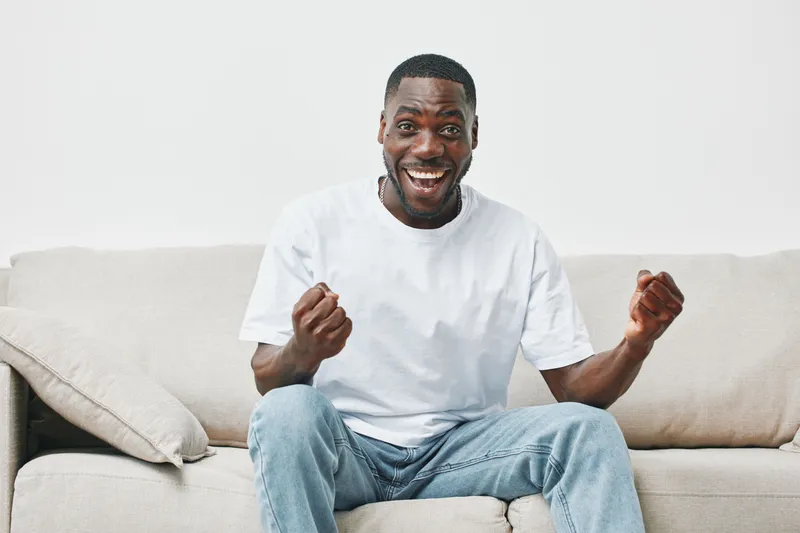 Happy young African American man celebrating on the sofa in a bright living room, wearing casual white t shirt and jeans, conveying joy and excitement