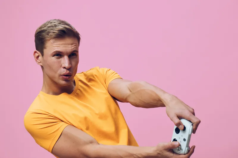 young caucasian man working with game controller in orange tshirt on pink background focused serious expression digital detox concept