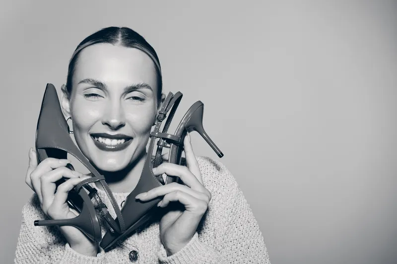 Smiling white woman with pale skin holds black high heel shoes near face wearing knitted sweater isolated on gray background. Concept for fashion and happiness.
