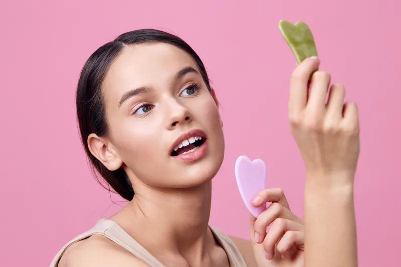 Young woman using facial massage tools against a pink background, showcasing skincare routine, beauty treatment, and self care concept with a joyful expression