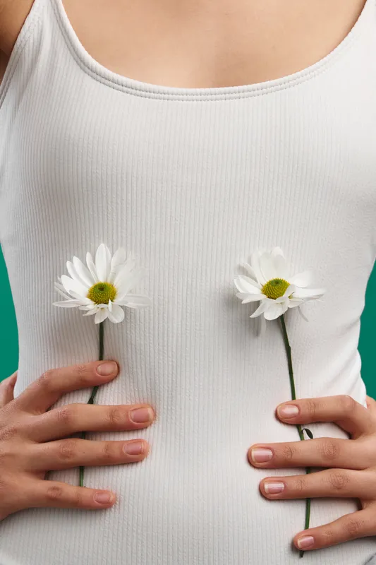 Asian woman wearing a white sleeveless top holds two daisies near her torso, creating a fresh, delicate moment with soft studio lighting and a clean, minimal background.