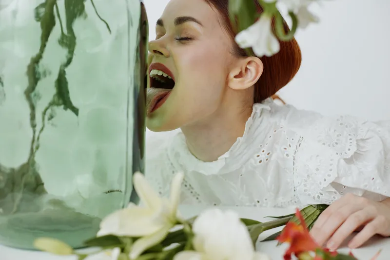A young model with red hair in a white lace blouse playfully licking a large glass vase surrounded by fresh flowers, conveying a sense of whimsy and innocence