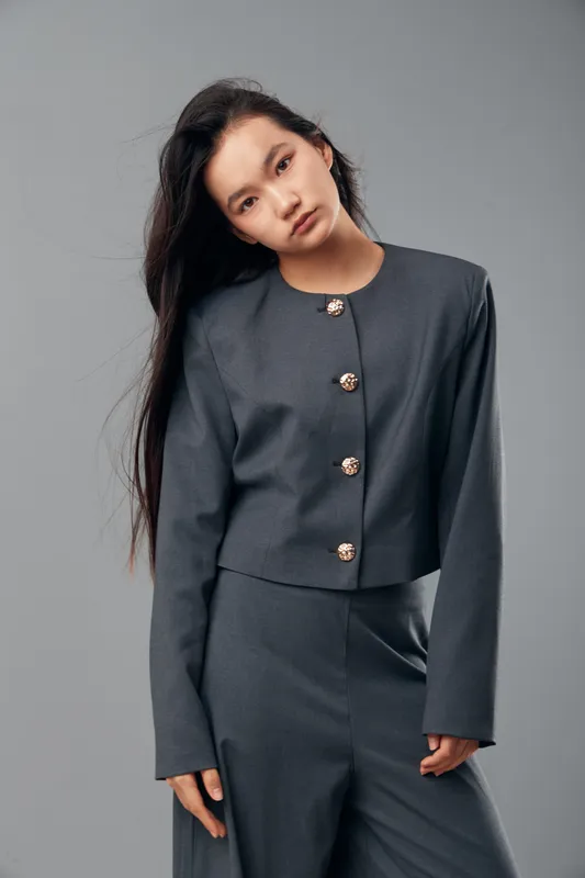 Young woman with long dark hair wearing gray suit jacket and matching pants posing against gray background. Fashion model with minimal makeup and serious expression in studio.