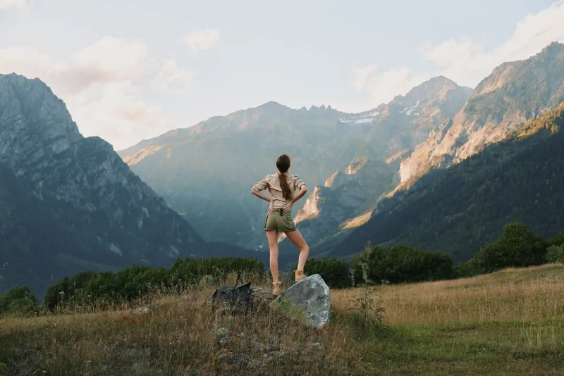 Woman stands on a rock in a mountain meadow, facing distant peaks, wearing outdoor gear, capturing a moment of adventure and solitude in nature amid scenic landscapes at dawn