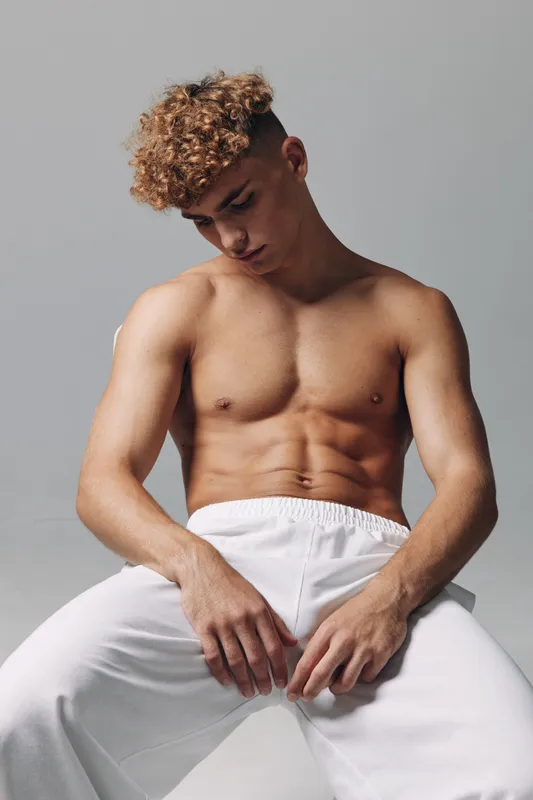 Young man with curly hair and muscular torso sitting wearing white pants, looking down in studio. Fit attractive player with pores microdetail, isolation and neutral background.