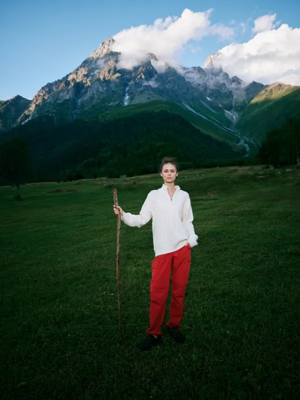 Mountain meadow scene with a woman in white sweater and red pants holding a staff, standing on grassy slope under cloudy sky, celebrating nature and rugged alpine landscape
