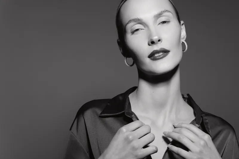Portrait of a slender Caucasian woman with fair skin and slicked-back hair wearing hoop earrings and a dark satin shirt. Concept of elegance, fashion, and minimalism in black and white.