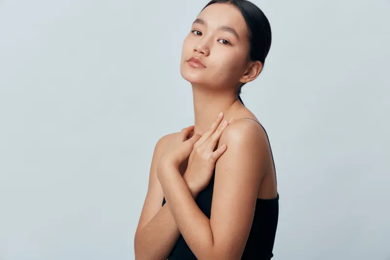 Young woman with smooth skin and dark hair posing with crossed arms in black sleeveless top on gray background for beauty, skincare, health and portrait concept.