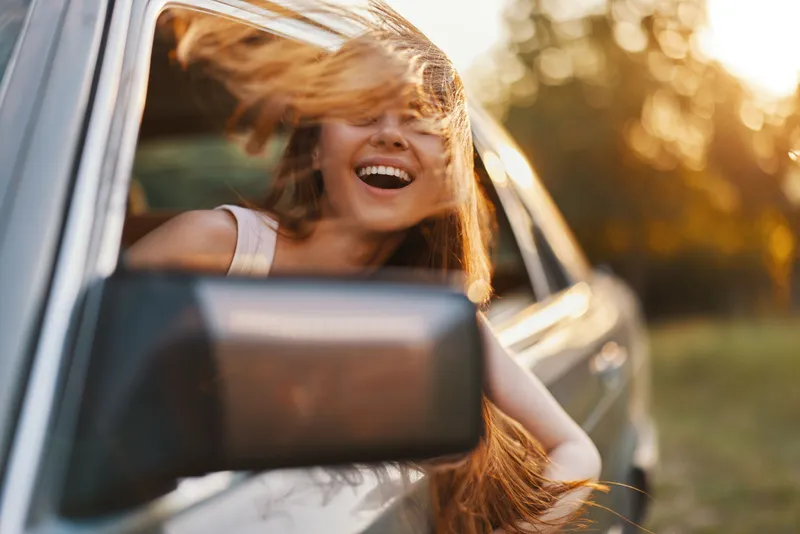 Happy woman enjoying a carefree drive in a car with flowing hair, sunlight shining through a blurred natural background, suggesting freedom and adventure.