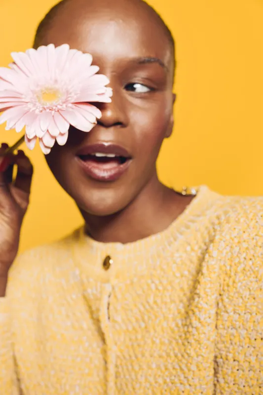 Close-up of a smiling black woman with dark skin holding a pink flower near her eye. She wears a yellow sweater and poses against a bright yellow background. Concept of beauty and joy.