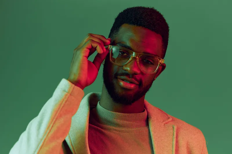 Young Black man in stylish glasses, smiling in a modern outfit against a green background, showcasing confidence and trendy fashion