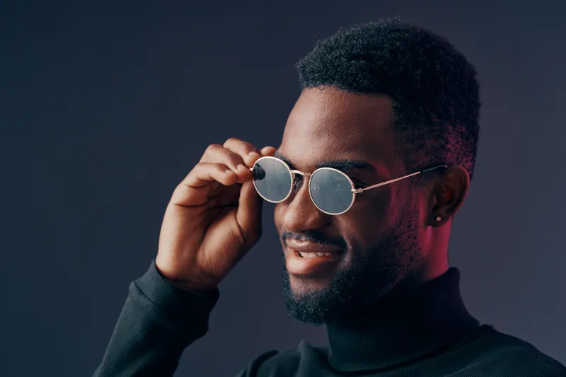 Stylish young Black man wearing sunglasses, smiling against a gradient background, showcasing modern fashion and vibrant colors in a portrait