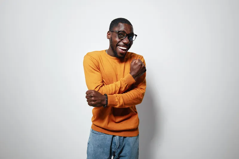 African American man with dark skin wearing orange sweater and glasses smiling and posing with crossed arms on white background. Casual style and happy expression.