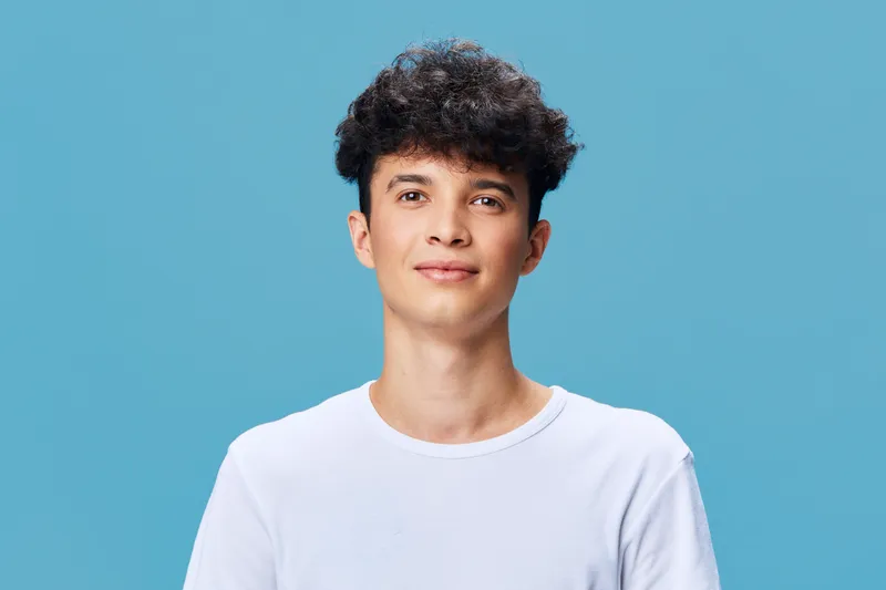 young man with curly hair wearing white shirt standing against blue background, looking confident and relaxed with neutral expression