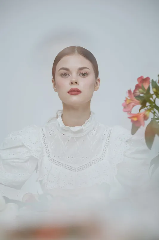 Elegant portrait of a young woman in a white lace dress with soft red lips, surrounded by fresh flowers against a light background