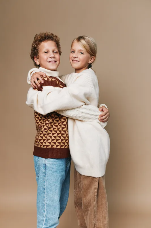Two children hug tightly in woolen sweaters, smiling at the camera. Warm tones, cozy knitwear, casual denim and corduroy create a heartwarming moment of friendship and comfort.