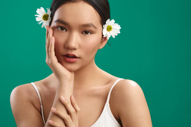 Asian woman with daisies tucked in her hair rests her face on her hand, posing softly against a vibrant green background. This serene portrait conveys natural beauty and calm confidence.
