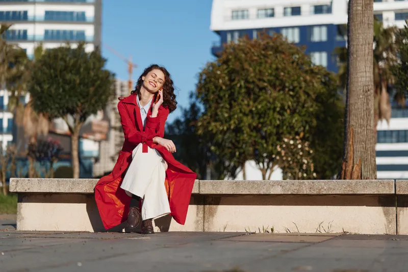 Young woman with curly hair wearing a red coat sits on a bench outdoors and talks on her phone with a joyful smile on a sunny day in the city.