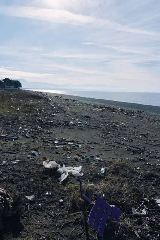 Polluted beach scene with plastic trash scattered along the shore, dark sand, cloudy sky, and a discarded chair piece, illustrating environmental damage from coastal litter and the need for cleaner