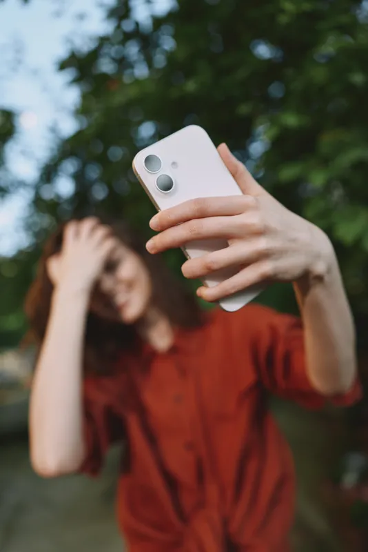 A woman lifestyle selfie outdoors, holding a phone in a casual outfit, bright natural light, smiling as she captures a carefree moment in nature, showcasing relatable self expression