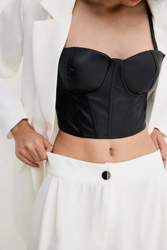 Stylish woman wearing black leather bustier and white tailored jacket adjusting high-waisted pants, showcasing modern fashion with minimalist chic and sleek design details.