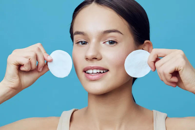 Young woman demonstrating skincare routine with cotton pads, smiling confidently against a bright blue background, showcasing healthy skin and wellness