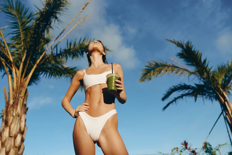 Bright midday scene shows a woman in white bikini holding a green smoothie on a sunny beach, palm trees frame the shot, casual lifestyle moment and carefree summer vibe.