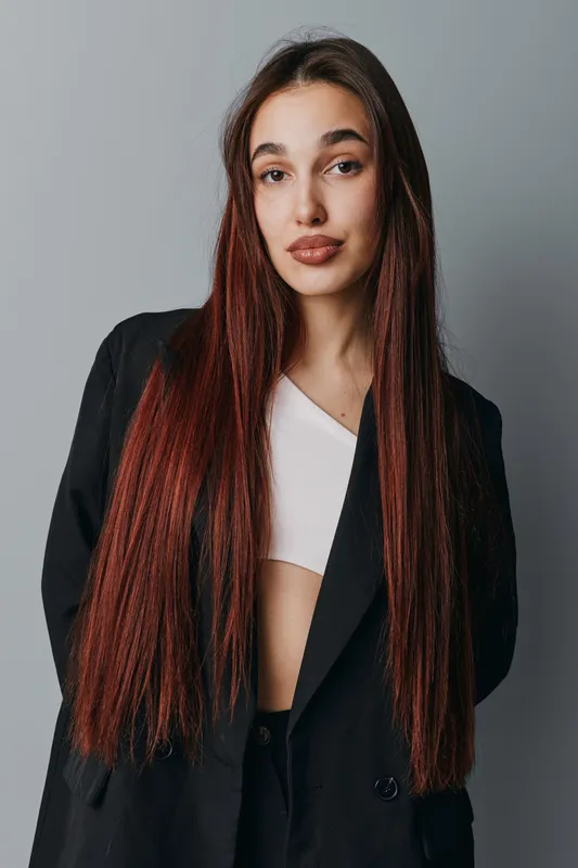 Woman with long red hair wearing black blazer and white top posing in studio with neutral background. Fashion portrait of confident female model with natural makeup and relaxed expression.