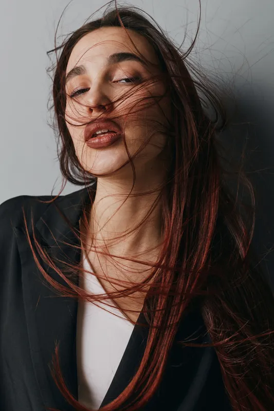 Woman with long hair blowing in wind wearing black blazer and white top posing against gray background with closed eyes and natural makeup for beauty and fashion portrait.