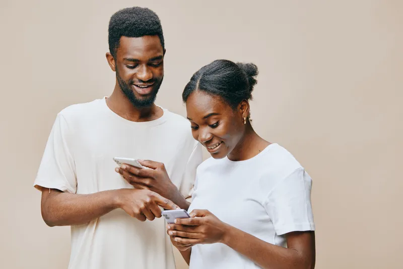 Happy Black couple smiling while using smartphones, showcasing connection and technology against a neutral background