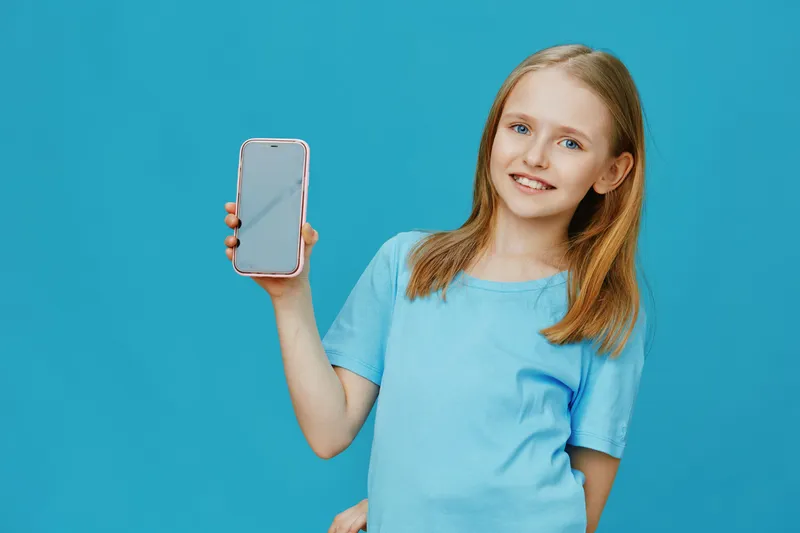 Girl smiling and holding smartphone against a bright blue background, showcasing modern technology and youthful enthusiasm