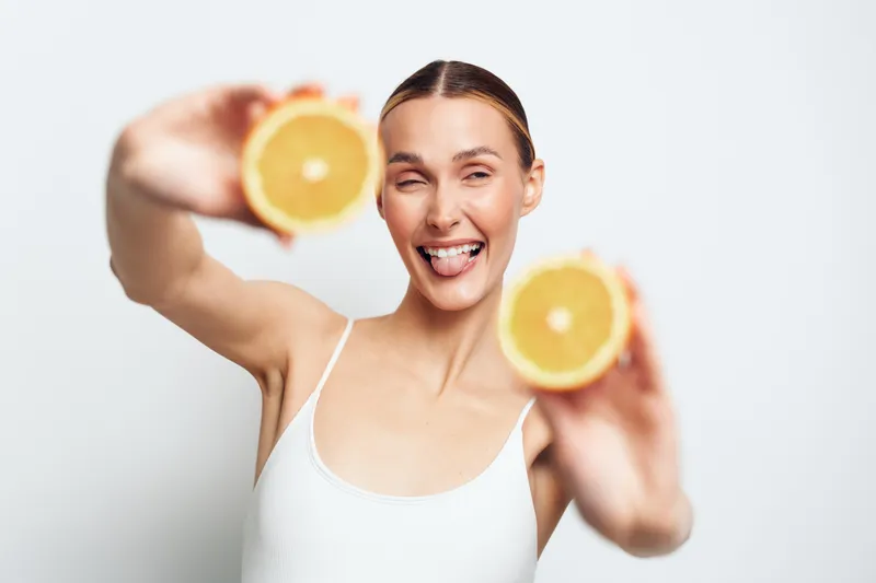 Playful woman holding two orange halves in front of her face, sticking out tongue, white tank top, bright studio background, fresh and fun mood.