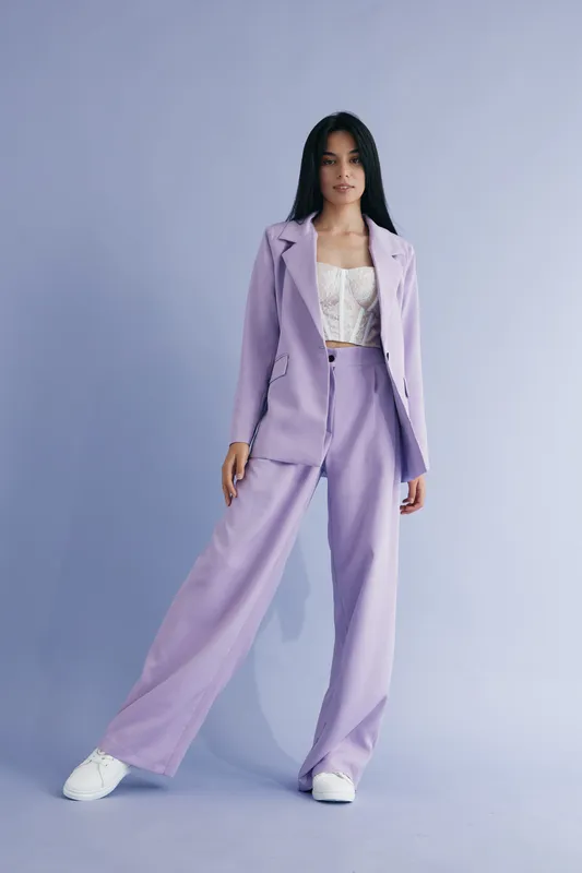 woman in lilac pantsuit stands confidently against pastel purple background wearing white lace top and sneakers, showcasing modern fashion and casual elegance