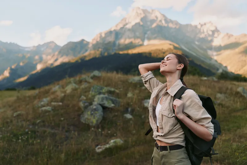 Cheerful woman hiking outdoors, backpack on, sunlit mountain landscape, relaxed posture, gentle breeze, vivid wildflowers, natural colors, vast scenery, wide sky above