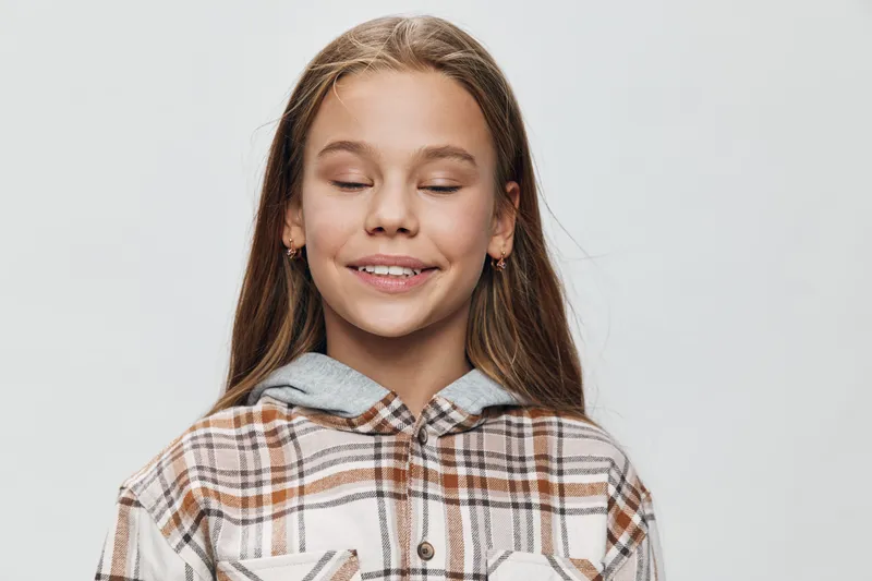 Young girl with closed eyes and smile wearing plaid shirt and earrings, portrait on light background. Child with long hair and calm expression for childhood and happiness.