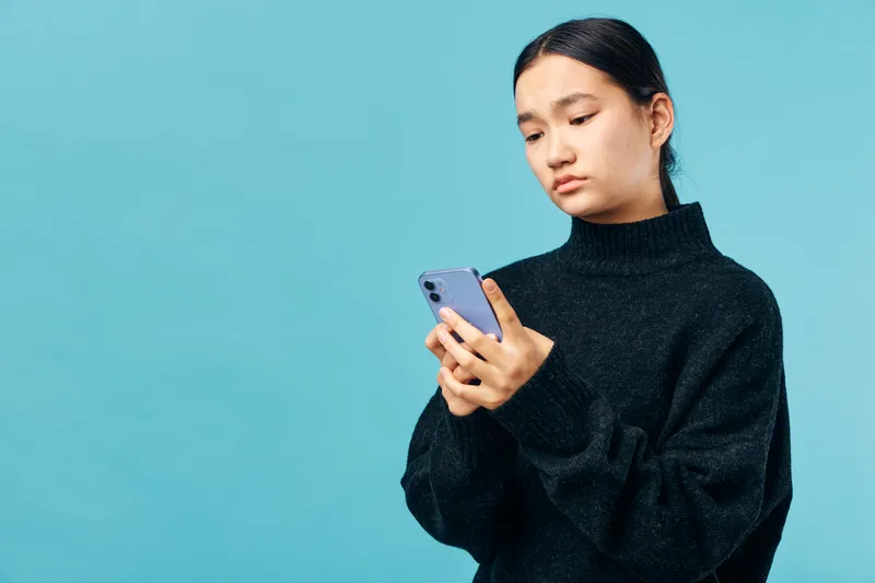 Young woman with dark hair in black sweater looking at smartphone, concentrating on screen with neutral expression, isolated on blue background for technology and communication concept.