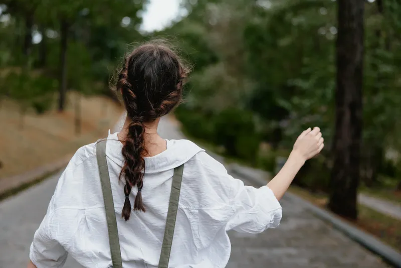 Young woman in casual white outfit walking outdoors, showcasing a relaxed vibe with a sense of freedom, surrounded by green nature on a quiet path