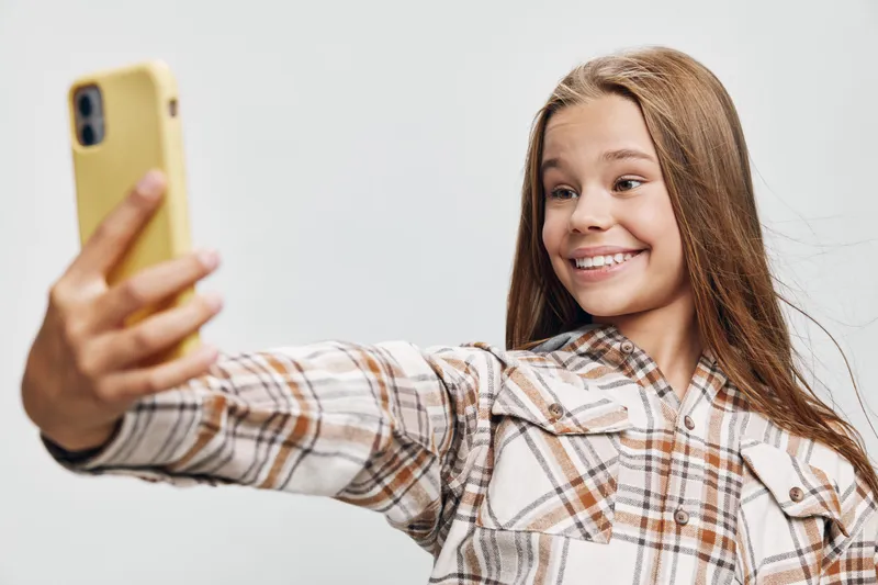 Girl taking selfie with smartphone smiling and looking at camera indoors, wearing plaid shirt. Happy teenager with long hair posing for photo in casual clothes on light background.