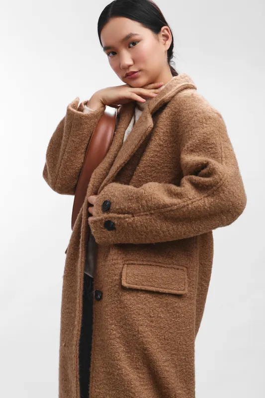 asian woman wearing a warm brown coat poses in a studio, gazing softly at the camera with a calm, confident demeanor that conveys modern style and quiet elegance.