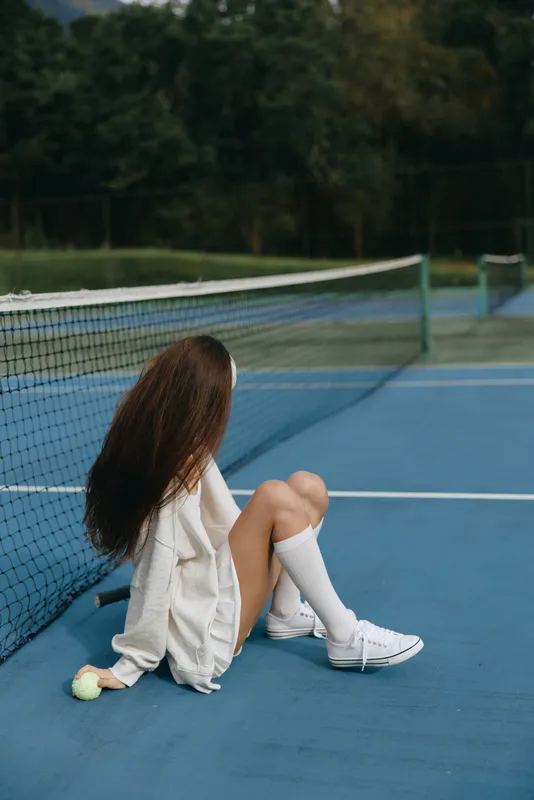 Young woman sitting on a tennis court, wearing casual sportswear, with long hair and a relaxed expression, surrounded by nature in a serene environment