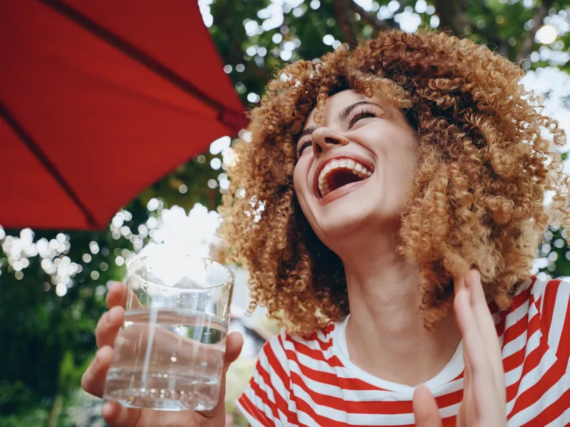 Joyful woman with curly hair smiles broadly while lifting a glass of water outdoors, under a vivid red umbrella, capturing a moment of refreshment and carefree laughter.