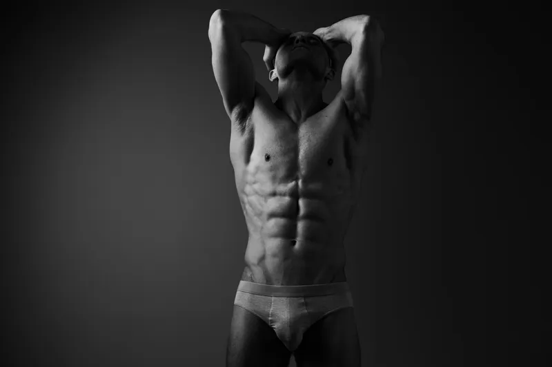 Black and white image of a muscular man posing confidently, showcasing strength and determination in a dramatic lighting setup.