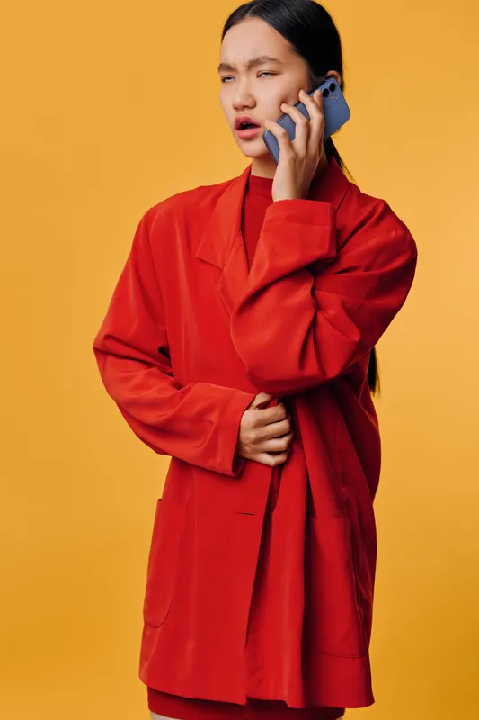 Woman in red jacket talking on mobile phone with confused expression on yellow background. Casual style, communication, connection, and emotion with modern technology, portrait and device.