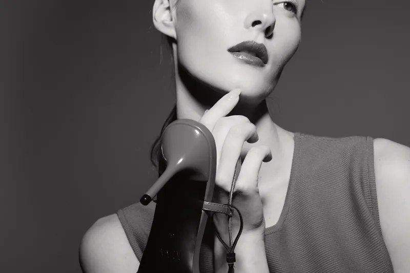 White woman with pale skin holds high heel shoe near her face in black and white studio portrait. Concept for fashion, elegance, and style with vintage mood and dramatic lighting.