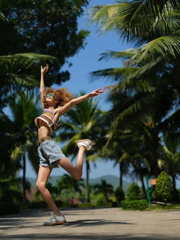 Energetic woman jumping outdoors in summer attire with vibrant blue sky and tropical palm trees in background, expressing joy and freedom.