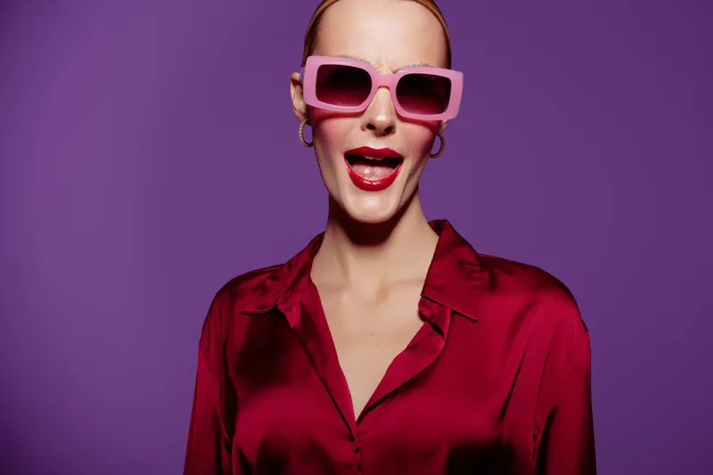 Stylish woman in red satin blouse and pink sunglasses poses confidently against purple background. Fashion portrait with bold colors and modern accessories for trendy style concept.