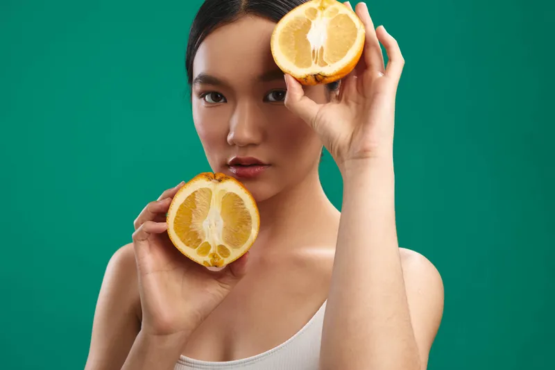 Asian woman holds fresh lemon halves against a vibrant green backdrop, presenting a bright, healthy mood with a zesty vibe and modern, inviting appeal.