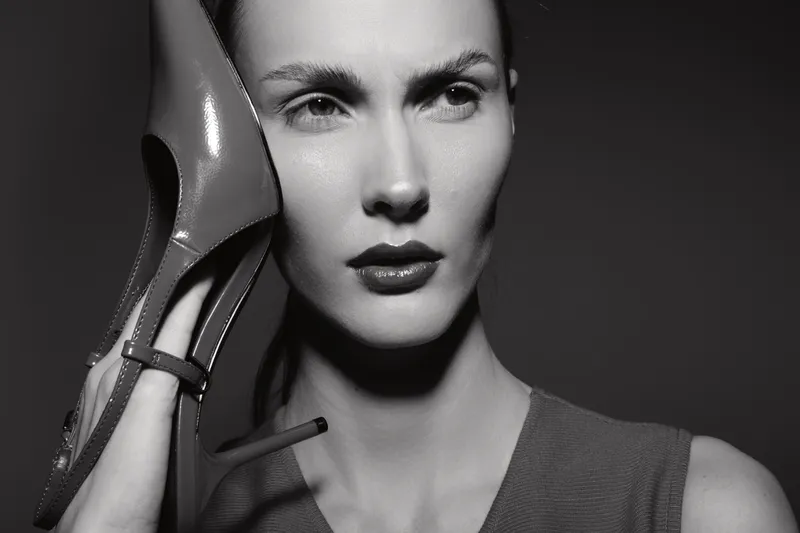 Close-up portrait of a serious white woman with pale skin holding a high heel shoe near her face. Studio lighting highlights facial features and creates dramatic shadows. Concept of fashion.