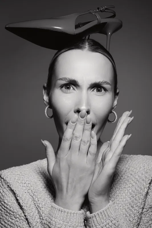 Portrait of a white woman with light skin covering her mouth with hands. A high heel shoe is balanced on her head. Concept for surprise and fashion creativity.