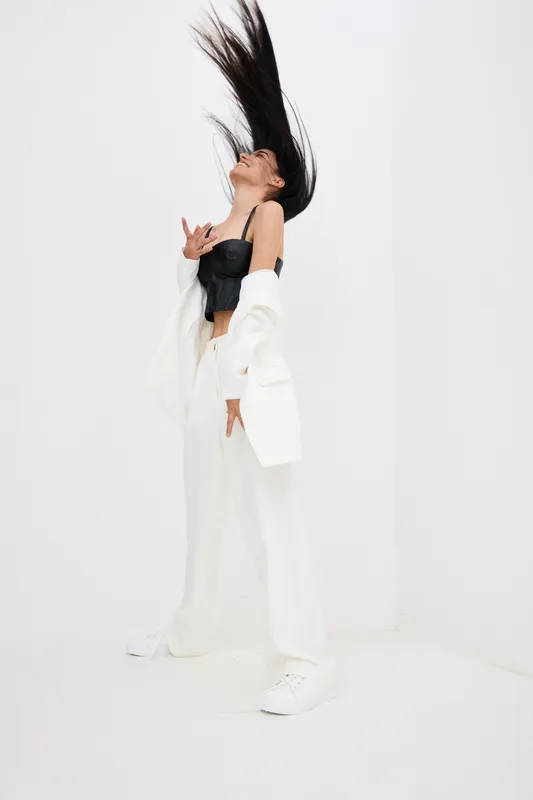 young woman in black top and white pants with dynamic hair movement posing against white wall, modern style, energetic and fashionable look